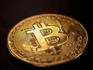 A close-up photograph of a physical gold-colored Bitcoin coin on a dark surface.