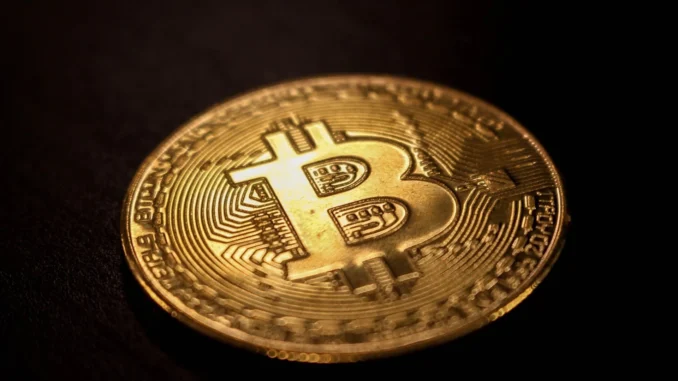A close-up photograph of a physical gold-colored Bitcoin coin on a dark surface.