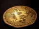 A close-up photograph of a physical gold-colored Bitcoin coin on a dark surface.